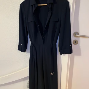 Svart jumpsuit från M&S Collection - Stilren svart långklänning från M&S Collection med knappar framtill, krage och trekvartsärmar. Klänningen har två bröstfickor, bälte i midjan och är tillverkad i ett mjukt, lätt material som ger en snygg siluett.