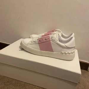 Valentino Garavani sneakers med rosa detalj - Säljer ett par vita Valentino Garavani sneakers med bred rosa rem över mitten och vita nitar på hälen. Skorna har rund tå och platt sula, samt klassisk snörning. Perfekta för dig som vill ha en stilren men ändå unik look.