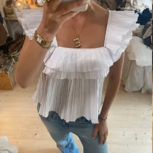 Vit plisserad blus med volang - Superfin vit blus med plisserat tyg och breda volangdetaljer över axlar och byst. Blusen har fyrkantig ringning och är ärmlös, perfekt till jeans eller kjol.