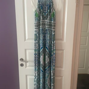 Mönstrad långklänning med halterneck - Säljer en långklänning med mönster i blått, grönt, svart och vitt. Klänningen har smala axelband och ett bohemiskt, paisleyinspirerat tryck. Perfekt för varma dagar och somriga tillfällen.