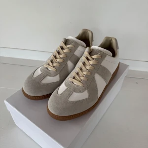 Maison Margiela gats - Säljer ett par Maison Margiela gats i vitt och beige med klassisk gummisula. Skorna har snörning och är tillverkade i en kombination av skinn och mocka. Perfekta för dig som gillar stilrena och tidlösa sneakers. Pris är diskuterbart.