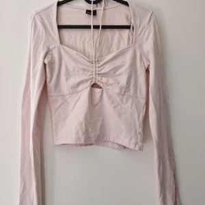 Ljusrosa långärmad croppad topp med knyt - Säljer en ljusrosa långärmad croppad topp med snygg rynkdetalj och liten cut-out framtill. Toppen har knytband vid halsen och är tillverkad i mjuk bomull. Perfekt för dig som gillar trendiga och feminina plagg.
