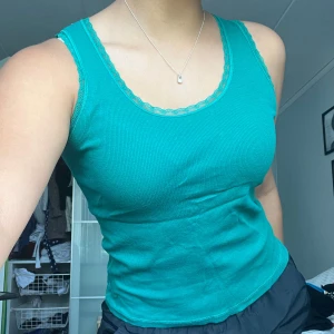 Turkos tanktop - Säljer en turkos tanktop med spetskant vid halsringningen. Toppen har en tight passform och är ärmlös, perfekt för varma dagar.