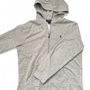 Grå hoodie med dragkedja från Polo Ralph Lauren - Klassisk grå hoodie från Polo Ralph Lauren med dragkedja framtill och huva. Tröjan är i nyskick och är i storlek L. Mitt pris 549 men kan diskuteras vid snabb affär! Skriv vid minsta fundering.