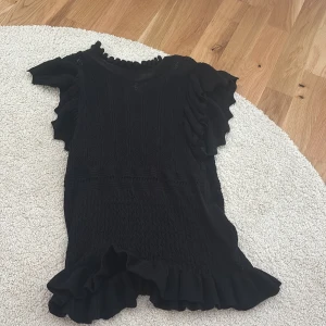 Svart volangklänning från Zara - Supersöt svart klänning från Zara med volangdetaljer vid ärmar och nederkant. Klänningen har korta ärmar och en feminin siluett som passar perfekt till många tillfällen. Stickad struktur och rund halsringning ger en trendig look.