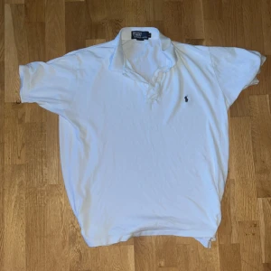 Vit pikétröja från Polo Ralph Lauren - Klassisk vit pikétröja från Polo Ralph Lauren med broderad logga på bröstet. Tröjan har korta ärmar, krage och knappar framtill. Perfekt för en stilren och tidlös look.