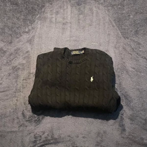 Svart stickad tröja från Polo Ralph Lauren - Klassisk svart stickad tröja från Polo Ralph Lauren med kabelstickat mönster och broderad logga på bröstet. Rund halsringning och långa ärmar. Perfekt för lager-på-lager-stil. 