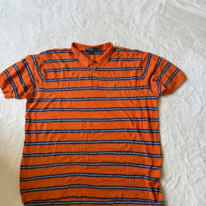 Orange randig pikétröja från Polo Ralph Lauren XXL - Orange pikétröja från Polo Ralph Lauren med blå och vita ränder. Klassisk krage och knäppning med två knappar. Tillverkad i 100% bomull och har en liten broderad logga på bröstet.