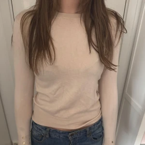 Beige långärmad tröja med gulddetaljer - Säljer en stilren beige långärmad tröja med rund halsringning. Tröjan har smal passform och dekorativa guldfärgade knappar vid ärmsluten. Perfekt att matcha med jeans eller kjol.