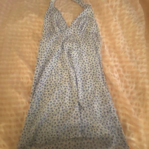 Blommig halterneckklänning från H&M - Söt somrig halterneckklänning från HM med blått blommönster på vit botten. Klänningen har omlott-v-ringning och knyts i nacken. Perfekt för varma dagar och har en lätt, luftig känsla.