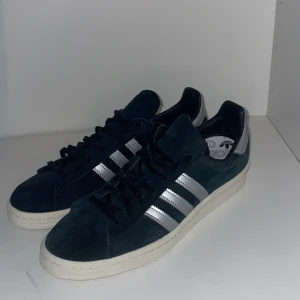 Adidas Campus sneakers med silvriga ränder - Mörkblå Adidas Campus sneakers i mocka med klassiska silvriga ränder på sidorna och vit sula. Skorna har svarta skosnören och silvriga detaljer vid hälen. Perfekta för en stilren vardagslook.