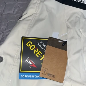 Vit och svart Gore-Tex jacka från The North Face - Säljer en snygg vit och svart jacka från The North Face med Gore-Tex material. Jackan har huva med dragsko, tryckknappar och dragkedja framtill samt logga på bröstet och axeln. Perfekt för blåsiga och regniga dagar.