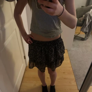 Svart blommig volangkjol - Supersöt svart kortkjol med små rosa blommor och volanger. Resår i midjan för skön passform. Ganska gammal kjol men inte använd så många gånger 🥰