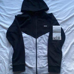 BOSS hoodie - Säljer en svart, grå och vit hoodie från BOSS med dragkedja framtill och huva. Tröjan har enkel design med BOSS-logga på bröstet och är tillverkad i mjukt material. Perfekt för en avslappnad stil. Bara att höra av sig om du har frågor