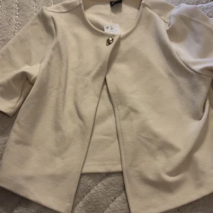 Beige kort kofta med knapp - Säljer en stilren beige koftan med trekvartsärm och en dekorativ knapp framtill. Koftan har en öppen design utan krage och är tillverkad i ett mjukt material som ger en elegant känsla.