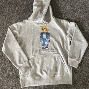 Grå hoodie med Polo Bear från Ralph Lauren - Säljer en grå hoodie från Ralph Lauren med den ikoniska Polo Bear-trycket framtill. Tröjan har huva med dragsko och en stor magficka. HELT NY🤩💫💫 (priset kan diskuteras)