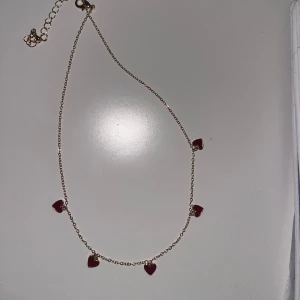 Guldfärgat halsband med röda hjärtan - Säljer ett tunt guldfärgat halsband med fem små röda hjärtan som hängande berlocker. Kedjan är justerbar och smycket har en söt och romantisk stil. Perfekt för att ge en färgklick till din outfit.