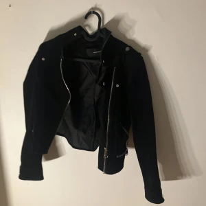 Svart skinnjacka från Vero Moda - Säljer en svart skinnjacka från Vero Moda med silvriga dragkedjor och klassisk bikerstil. Jackan har långa ärmar och flera detaljer som snedställd dragkedja och fickor med dragkedja. Perfekt för en cool och stilren look.
