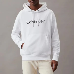 Vit hoodie från Calvin Klein - Säljer en stilren vit hoodie från Calvin Klein med svart logotyp på bröstet. Tröjan har huva med dragsko och en stor magficka. Perfekt för en avslappnad och trendig look.