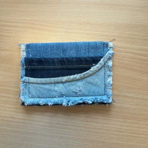 Plånbok i jeans med råa kanter - Säljer en unik plånbok sydd av återvunnet jeanstyg i olika blå nyanser. Plånboken har råa, fransiga kanter och flera fack på framsidan för kort eller småsaker. Perfekt för dig som gillar en personlig och kreativ stil!