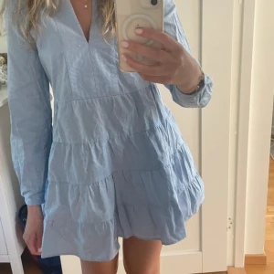 Ljusblå volangklänning med lång ärm - Supersöt ljusblå klänning med volanger och v-ringning. Klänningen har långa ärmar och en avslappnad passform med flera volanglager. Perfekt för vår och sommar, och lätt att styla med sneakers eller sandaler.