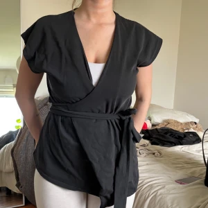 Svart omlottopp med knytband från Zara - Säljer en svart omlottopp från Zara med korta ärmar och knytband i midjan. Toppen har v-ringning och en mjuk, följsam passform som ger en snygg siluett. Man kan knyta den på olika sätt som man ser på bilderna, aldrig använt! Helt ny 🩷 