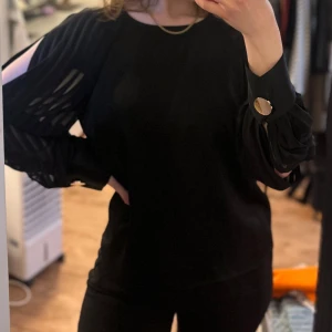 Svart blus med vida ärmar och guldknappar - Säljer en svart blus med vida, plisserade ärmar och snygga slitsar längs ärmarna. Blusen har stora guldiga knappar vid ärmsluten som en extra detalj. Perfekt för dig som gillar stilrena plagg med en twist.