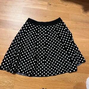 Svart polkadot kjol, prickig kjol - Säljer en polkadot kjol, svart kjol med vita prickar. Kjolen har en utställd form och resår i midjan för extra komfort. Perfekt för dig som gillar klassiska mönster och vill ha en lekfull touch i garderoben. Nästan aldrig använt, i jättebra skick!