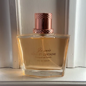 Je suis Sûre et Certaine Bouquet de Fleurs Eau de Parfum - Elegant parfymflaska med roséguldfärgat lock och transparent glas. Doften heter Sûre et Certaine Bouquet de Fleurs och är en Eau de Parfum. Flaskan har en modern, kantig form och rymmer en ljusgul vätska.