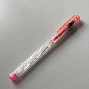Genomskinlig bläckpenna med rosa och orange detaljer - Säljer en genomskinlig bläckpenna med rosa spets och orange lock. Pennan har en smal och lätt design, perfekt för anteckningar eller skolarbete. Bläckpatronen syns tydligt genom pennkroppen.