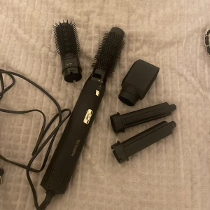 Cosmopolitan 5-in-1 Hot Air Styling Set - Multifunktionell styling set med 5 utbytbara tillbehör för hårstyling. Inkluderar rundborste, volymborste och olika munstycken. Perfekt för att skapa volym, lockar eller släta frisyrer. Svart design med gulddetaljer och roterbar sladd.
