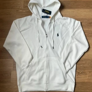 Vit hoodie från Polo Ralph Lauren - Säljer en klassisk vit hoodie från Polo Ralph Lauren med dragkedja och huva. Tröjan har det ikoniska broderade logotypen på bröstet och praktiska fickor framtill. Perfekt för en stilren och avslappnad look.