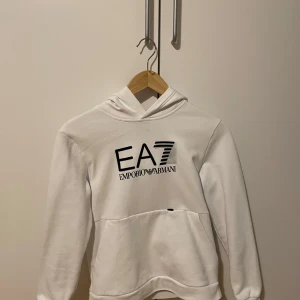 Vit hoodie från Emporio Armani EA7 - Säljer en stilren vit hoodie från Emporio Armani EA7 med svart logga framtill. Tröjan har huva och en stor magficka. Perfekt för en avslappnad och trendig look.