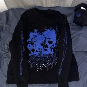 Svart långärmad tröja med blått dödskalletryck - Säljer en svart långärmad tröja med stort blått dödskalletryck framtill och taggtrådsmönster längs ärmarna. Tröjan har rund hals och är i mjukt material, perfekt för en streetstil.