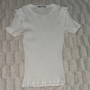 Vit ribbad topp med volangärm från Zara - Säljer en söt vit ribbad topp från Zara med korta ärmar och volangdetalj vid axlarna. Perfekt till sommaren och enkel att matcha med jeans eller kjol.