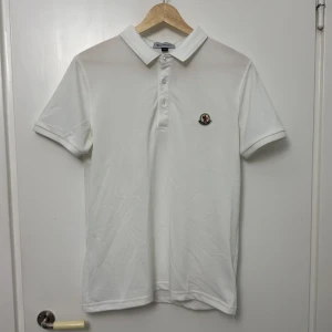 Vit piké t-shirt från Moncler - Säljer en klassisk vit piké t-shirt från Moncler med liten broderad logga på bröstet. T-shirten har krage, knappar framtill och är kortärmad. Perfekt för en clean och stilren look.