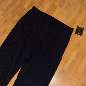 Mörkblå randiga kostymbyxor från GR Design - Snygga mörkblå kostymbyxor med tunna vita ränder från GR Design. Byxorna har hög midja, raka ben och stängs med dragkedja och knappar. Perfekta för en stilren look och har bälteshällor för extra detalj.