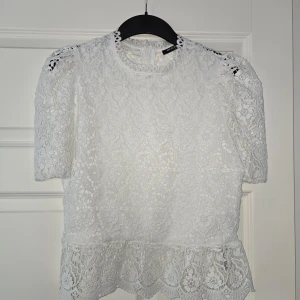 Vit spetsblus med volang - Säljer en romantisk vit spetsblus med korta puffärmar och volang nertill. Blusen har vackra blommiga spetsdetaljer och rund halsringning. Perfekt för dig som gillar en feminin och trendig stil.