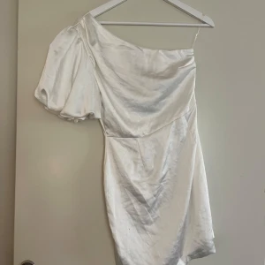 Vit satin klänning med puffärm och oneshoulder - Säljer en snygg vit kortklänning i satin med en trendig oneshoulder-design och puffärm. Oanvänd med prislapp kvar. Strl 36