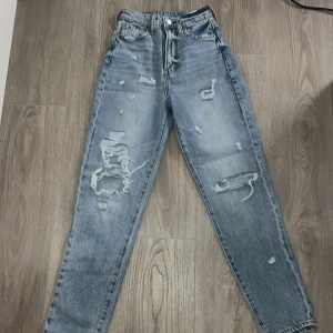 Blå mom jeans med slitningar från H&M - Säljer ett par blå mom jeans från H&M med hög midja och raka ben. Jeansen har flera slitningar och hål framtill för en trendig look. Klassisk femficksmodell och knappgylf. Perfekta till vardagsstilen!