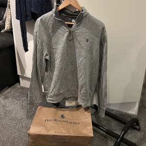 Grå halvzip tröja från Polo Ralph Lauren - Säljer en klassisk grå halvzip tröja från Polo Ralph Lauren. Tröjan har lång ärm, liten broderad logga på bröstet och hög krage med dragkedja. Perfekt för en stilren och avslappnad look.