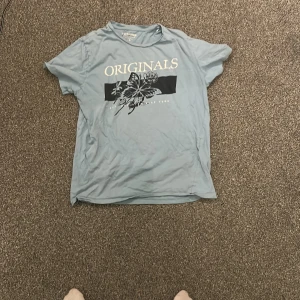Ljusblå t-shirt med tryck från Jack & Jones - Säljer en ljusblå t-shirt från Jack & Jones med texten 'ORIGINALS' och ett blommotiv på bröstet. T-shirten har rund hals och korta ärmar. Perfekt för en avslappnad stil. Det finns tyvärr en fläck på ryggen som säkert går bort jag har bara inte testat men vid intresse av tröjan så fixar jag