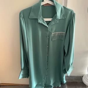 Mintgrön skjorta med pärldetaljer - Snygg mintgrön skjorta med långa ärmar och klassisk krage. Skjortan har knappar framtill och en unik detalj med pärlor på bröstfickan. Perfekt för dig som vill sticka ut lite extra med en stilren touch.