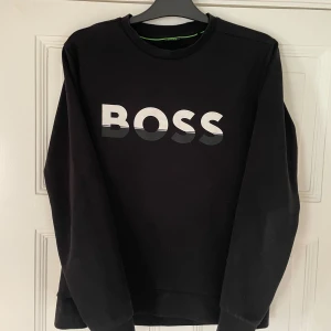 Svart sweatshirt från BOSS - Säljer en svart sweatshirt från BOSS med stor logga framtill i vitt och grått. Tröjan har rund halsringning och långa ärmar. Perfekt för en stilren och avslappnad look.