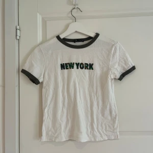 Vit t-shirt med New York-tryck från Bershka - Vit t-shirt från Bershka med grönt och svart 'NEW YORK'-tryck på bröstet. T-shirten har kontrastfärgade mörkgrå kanter vid halsringning och ärmslut. Klassisk passform och korta ärmar.