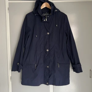 Mörkblå parkajacka från Isolde 40 - Mörkblå parkajacka från Isolde med dragkedja och knappar framtill. Jackan har en huva och praktiska fickor med dragkedja. Dragsko i midjan. Storlek 40