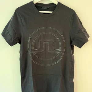 Svart t-shirt med logga från J.Lindeberg - Säljer en svart t-shirt från J.Lindeberg med stor, diskret logga på bröstet. Klassisk rund halsringning och normal passform. Perfekt för en stilren och enkel look.