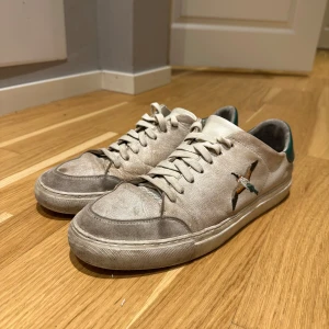 Axel Arigato Skor  - Säljer ett par vita sneakers med grå detaljer och broderad fågel på sidan. Skorna har snörning och rund tå, samt en grön detalj på hälen. 
