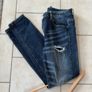 Mörkblå slitna jeansbyxor från dsquared - Säljer ett par mörkblå jeansbyxor från dsquared med slitningar och hål på framsidan. Klassisk femficksmodell med dragkedja och knapp. Tapered fit och snygga detaljer vid fickorna. Perfekta för en avslappnad stil.