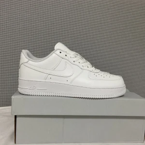 Nike Air Force 1 helvita sneakers - Klassiska helvita Nike Air Force 1 Low sneakers med snörning och perforerad tå. Defekt på skolådan men annars är skorna helt nya. Skorna har en platt sula och är tillverkade i syntetmaterial och skinn. Perfekta för dig som gillar en clean och tidlös stil.